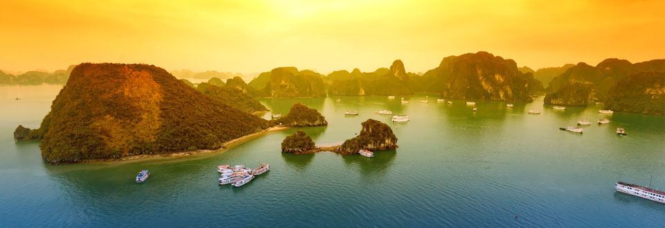 Dramatiskt solnedgångsljus över Halong Bay med kalkstensöar och segelbåtar.