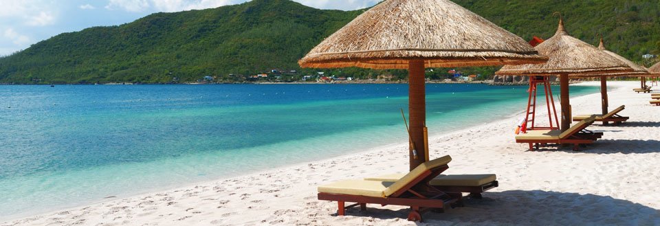 Vit sandstrand med strandparasoll och solstolar framför en klar turkos hav och gröna berg i bakgrunden.