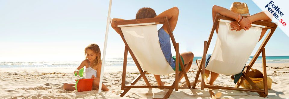 En familj njuter av solen på stranden, barnen leker i sanden medan vuxna vilar i solstolar.