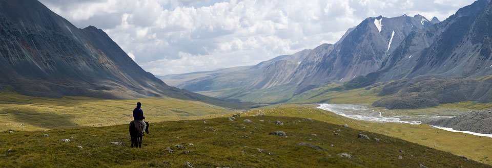 En ryttare på häst blickar över ett imponerande bergslandskap med öppna fält och en flod.